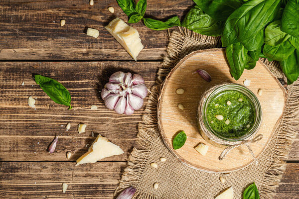 Homemade Italian basil pesto sauce in a glass jar. Fresh bunch of leaves, parmesan, pine nuts, and olive oil. Trendy hard light, dark shadow, old wooden background, top view