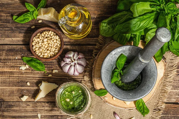 Homemade Italian basil pesto sauce in a vintage mortar with pestle. Fresh bunch of leaves, parmesan, pine nuts, and olive oil. Trendy hard light, dark shadow, old wooden background, top view