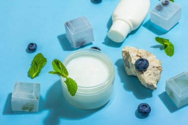 Natural skincare. Cream or lotion with organic berries. Blueberry, mint, and ice. Moisturizing and tonic cosmetics concept, flat lay, trendy hard light, dark shadow, blue background, close up