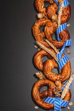 Oktoberfest concept - pretzels with traditional pattern ribbon and ears of wheat. A trendy hard light, dark shadow, black stone concrete background, flat lay, top view