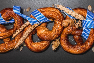 Oktoberfest concept - pretzels with traditional pattern ribbon and ears of wheat. A trendy hard light, dark shadow, black stone concrete background, flat lay, top view