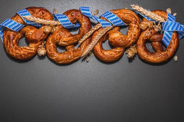 Oktoberfest concept - pretzels with traditional pattern ribbon and ears of wheat. A trendy hard light, dark shadow, black stone concrete background, flat lay, top view