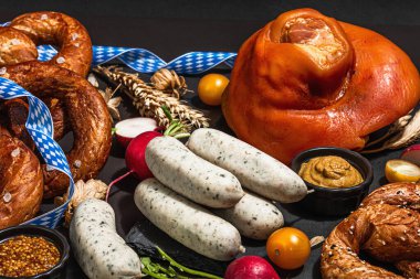 Traditional Oktoberfest set. Pretzels, weisswurst, smoked pork knuckle eisbein with hop. German festival food concept. Trendy hard light, dark shadow, black stone concrete background, close up
