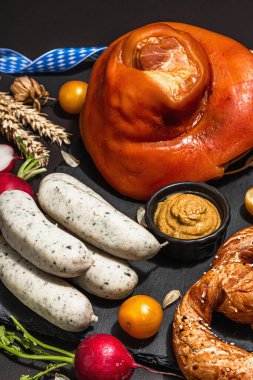 Traditional Oktoberfest set. Pretzels, weisswurst, smoked pork knuckle eisbein with hop. German festival food concept. Trendy hard light, dark shadow, black stone concrete background, close up