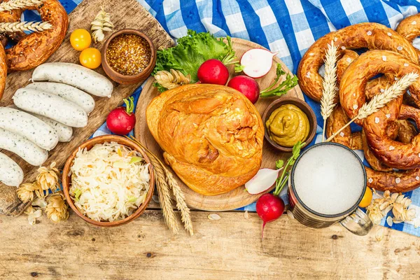 Traditional Oktoberfest set. Pretzels, beer, weisswurst, smoked pork knuckle eisbein with fermented cabbage and mustard. German festival food concept. Old wooden background, top view