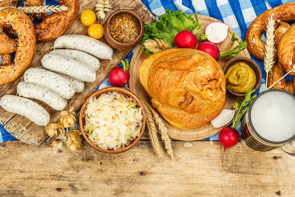 Traditional Oktoberfest set. Pretzels, beer, weisswurst, smoked pork knuckle eisbein with fermented cabbage and mustard. German festival food concept. Old wooden background, top view