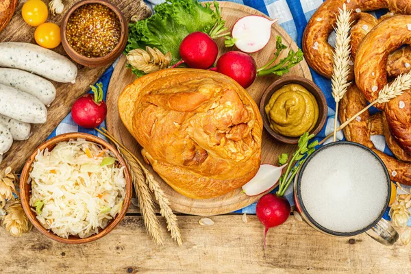 Traditional Oktoberfest set. Pretzels, beer, weisswurst, smoked pork knuckle eisbein with fermented cabbage and mustard. German festival food concept. Old wooden background, top view