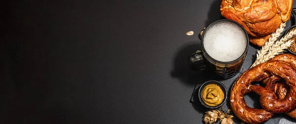 Traditional Oktoberfest set. Pretzels, beer, smoked pork knuckle eisbein with mustard. German festival food concept. Trendy hard light, dark shadow, black stone concrete background, banner format