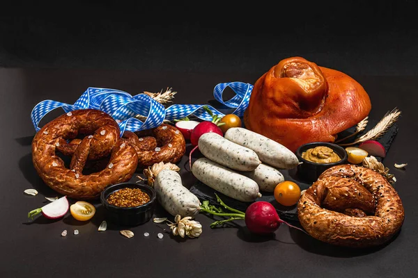Traditional Oktoberfest set. Pretzels, weisswurst, smoked pork knuckle eisbein with hop. German festival food concept. Trendy hard light, dark shadow, black stone concrete background, copy space