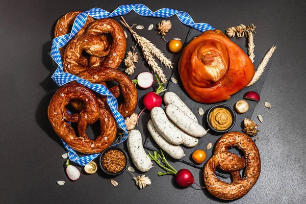 Traditional Oktoberfest set. Pretzels, weisswurst, smoked pork knuckle eisbein with hop. German festival food concept. Trendy hard light, dark shadow, black stone concrete background, top view