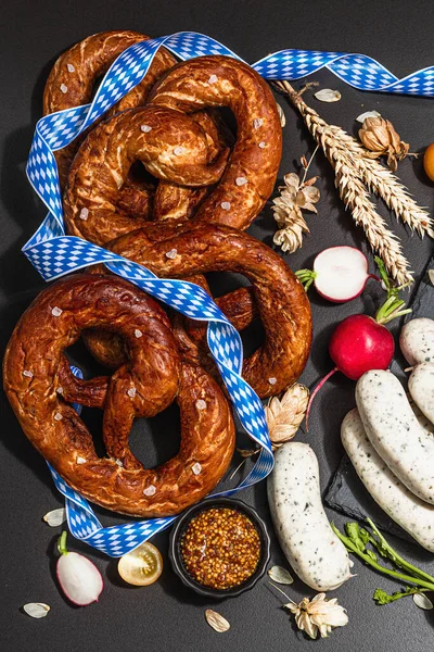 Traditional Oktoberfest set. Pretzels, weisswurst, smoked pork knuckle eisbein with hop. German festival food concept. Trendy hard light, dark shadow, black stone concrete background, top view