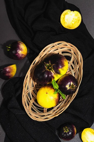 Yellow-violet Tomatoes variety Primary colors in a wicker basket. Harvest of ripe vegetables, trendy hard light, dark shadow. Black stone concrete background, top view