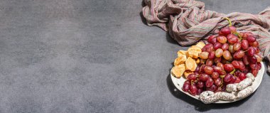 Peynirli abur cubur tabağı, üzüm ve kurutulmuş sosis. Şarküteri antipasto, taş beton arka plan. Modaya uygun sert ışık, koyu gölge, düz döşeme, afiş biçimi