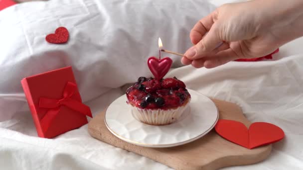 Hand zündet eine Kerze auf einer Torte auf dem Bett mit rotem Valentinstagsdekor und Herzen an
