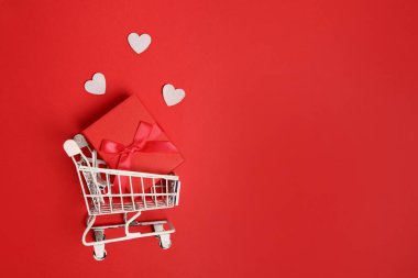 Hediye kutulu alışveriş arabası, kırmızı arka planda beyaz kalpler. Sevgililer Günü alışverişi ve satışı