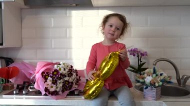 8 Mart, tatlı kız bebek balonla oynuyor ve evdeki çiçeklerle mutfak masasında oturuyor.