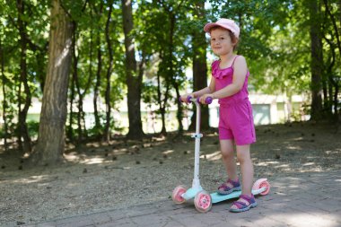 Pembe elbiseli kız parkta scooter sürüyor.