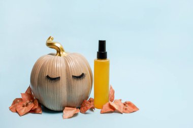 Cosmetic spray oil bottle and pumpkin with false eyelashes on blue background with autumn leaves, hair and facial skin care in autumn