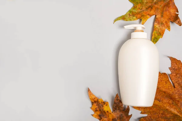 Soap or lotion bottle with dry autumn leaves on light background, autumn cosmetics