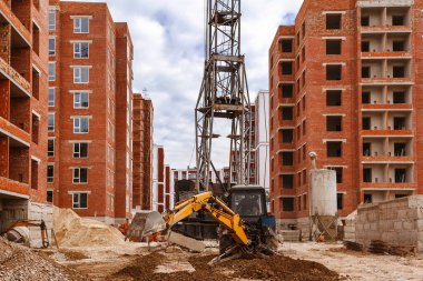 Kazıcı, kule vinci olan tuğla bir yapının arka planında bir inşaat alanında çalışıyor.