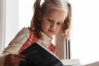 Tepe ve okuma. Pencerede kitap okuyan sevimli küçük bir kız. Aletsiz çocuk eğlencesi kavramı.