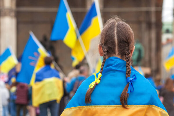 Rally support Ukraine in Europe. Ukraine Flag on Shoulders of Girl with blue yellow ribbons on flags background. Stand with Ukraine concept. 