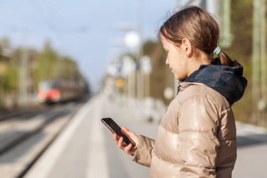 Demiryolu platformunda akıllı telefon kullanılıyor. Tren istasyonunda bekleyen bir kız akıllı telefona bakıyor. Online bilet rezervasyonu.