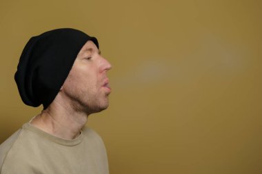 A young European lets off steam from an electronic cigarette. Portrait of a white man in a black cap.