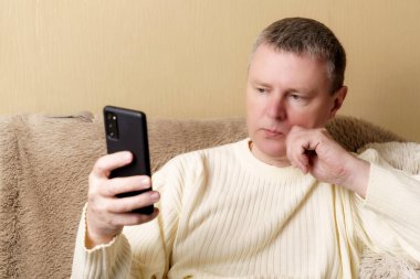 A European in a white sweater sits on a sofa with a phone in his hand. A man looks thoughtfully into a smartphone, resting his head on his hand