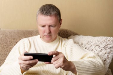 Haberleri okuyan ya da akıllı telefondan oynayan bir adam. Beyaz kazaklı bir Avrupalı elinde telefonla koltukta oturuyor..