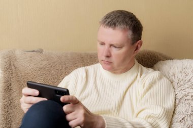 Beyaz kazaklı bir Avrupalı elinde telefonla kanepede oturuyor. Haberleri okuyan ya da akıllı telefondan oynayan adam.