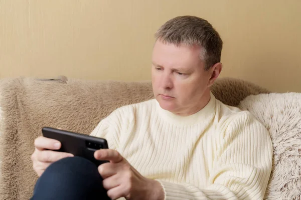 Beyaz kazaklı bir Avrupalı elinde telefonla kanepede oturuyor. Haberleri okuyan ya da akıllı telefondan oynayan adam.