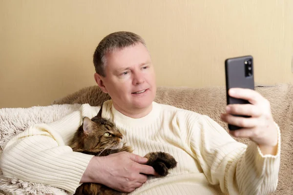 A man with a cat in his arms in a video chat with relatives or friends. A European in a white sweater with a phone in his hand sits on a sofa, holds a cat in his arms — стокове фото