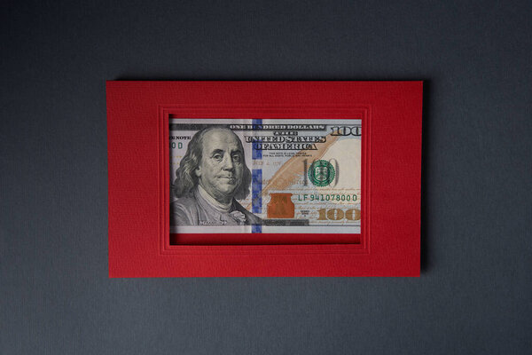 One hundred dollars banknote in a red cardboard frame on a gray background.