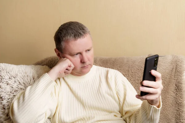 Bir adam akıllı bir telefona dikkatlice bakar, başını elinin üzerine koyar. Beyaz kazaklı bir Avrupalı elinde telefonla koltukta oturuyor..