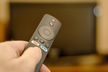 TV kumandası elinde, düğmelerde yazılar ve sahte haberler var. Kırmızı yazılar sahte, mavi yazılar ise uzaktan kumandanın düğmelerinde..