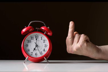Sabah uyanışı. Saat yedide güneş ışınlarında okları olan kırmızı alarm saati ve orta parmağı, koyu arkaplanı gösteren bir el..
