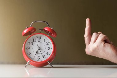 Sabah uyanışı. Saat yedide güneş ışınlarında okları olan kırmızı alarm saati ve orta parmağı gösteren bir el..