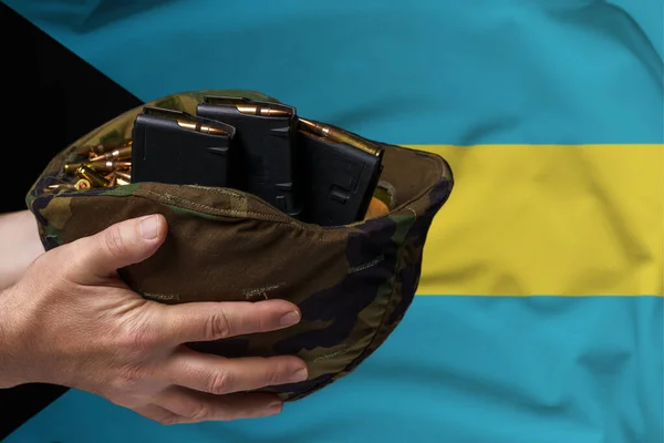 A military helmet with cartridges and magazines for a rifle in the hands of a man against the background of the flag of Bahamas. The concept of selling weapons or military assistance.