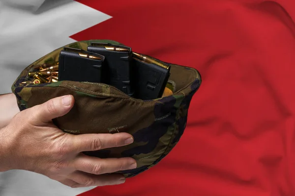 A military helmet with cartridges and magazines for a rifle in the hands of a man against the background of the flag of Bahrain. The concept of selling weapons or military assistance.
