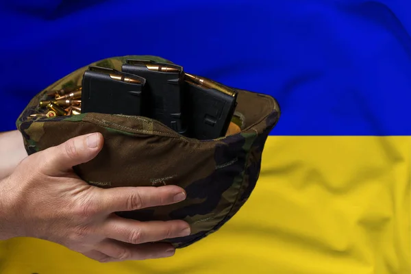 ウクライナの国旗を背景に男の手にライフルのためのカートリッジと雑誌を持つ軍事ヘルメット 武器の販売又は軍事援助の概念 — ストック写真