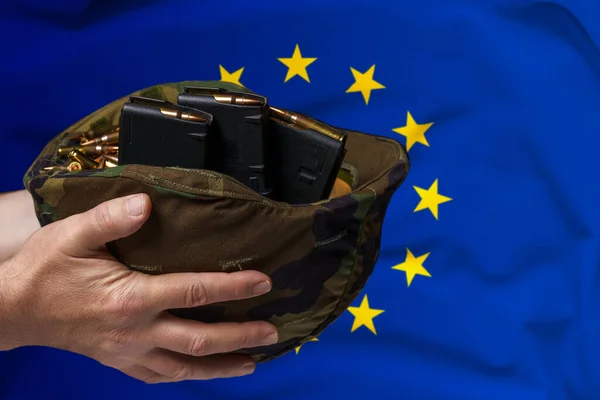 A military helmet with cartridges and magazines for a rifle in the hands of a man against the background of the flag of European Union. The concept of selling weapons or military assistance.