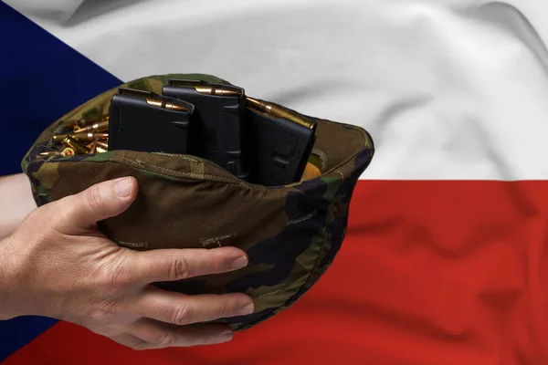 A military helmet with cartridges and magazines for a rifle in the hands of a man against the background of the flag of Czechia. The concept of selling weapons or military assistance.