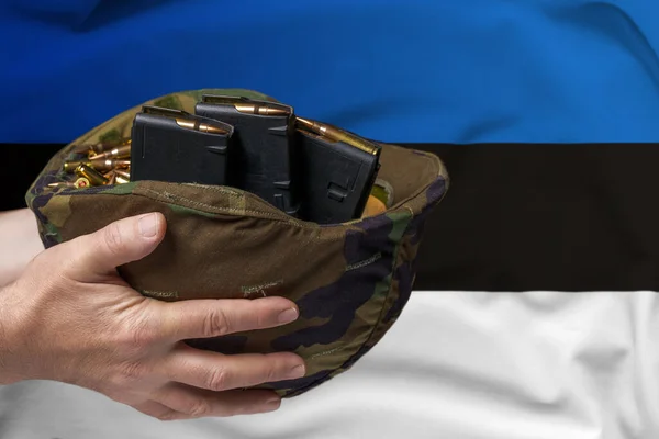 A military helmet with cartridges and magazines for a rifle in the hands of a man against the background of the flag of Estonia. The concept of selling weapons or military assistance.