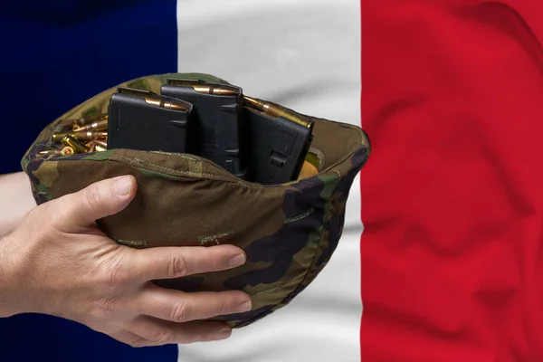 A military helmet with cartridges and magazines for a rifle in the hands of a man against the background of the flag of France. The concept of selling weapons or military assistance.