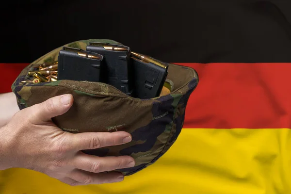 A military helmet with cartridges and magazines for a rifle in the hands of a man against the background of the flag of Germany. The concept of selling weapons or military assistance.