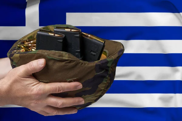 在希腊国旗的背景下 一个手拿着步枪弹夹和弹夹的男人手中的军事头盔 出售武器或军事援助的概念 — 图库照片