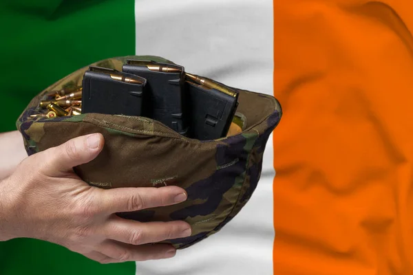 A military helmet with cartridges and magazines for a rifle in the hands of a man against the background of the flag of Ireland. The concept of selling weapons or military assistance.