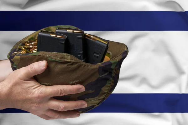 A military helmet with cartridges and magazines for a rifle in the hands of a man against the background of the flag of Israel. The concept of selling weapons or military assistance.
