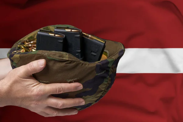 在拉脱维亚国旗的背景下 一个手握步枪弹夹和弹夹的男人手中的军事头盔 出售武器或军事援助的概念 — 图库照片
